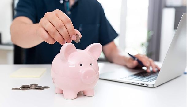 Ein Mensch wirft Geld in ein Sparschwein, während er vor einem Notebook sitzt.